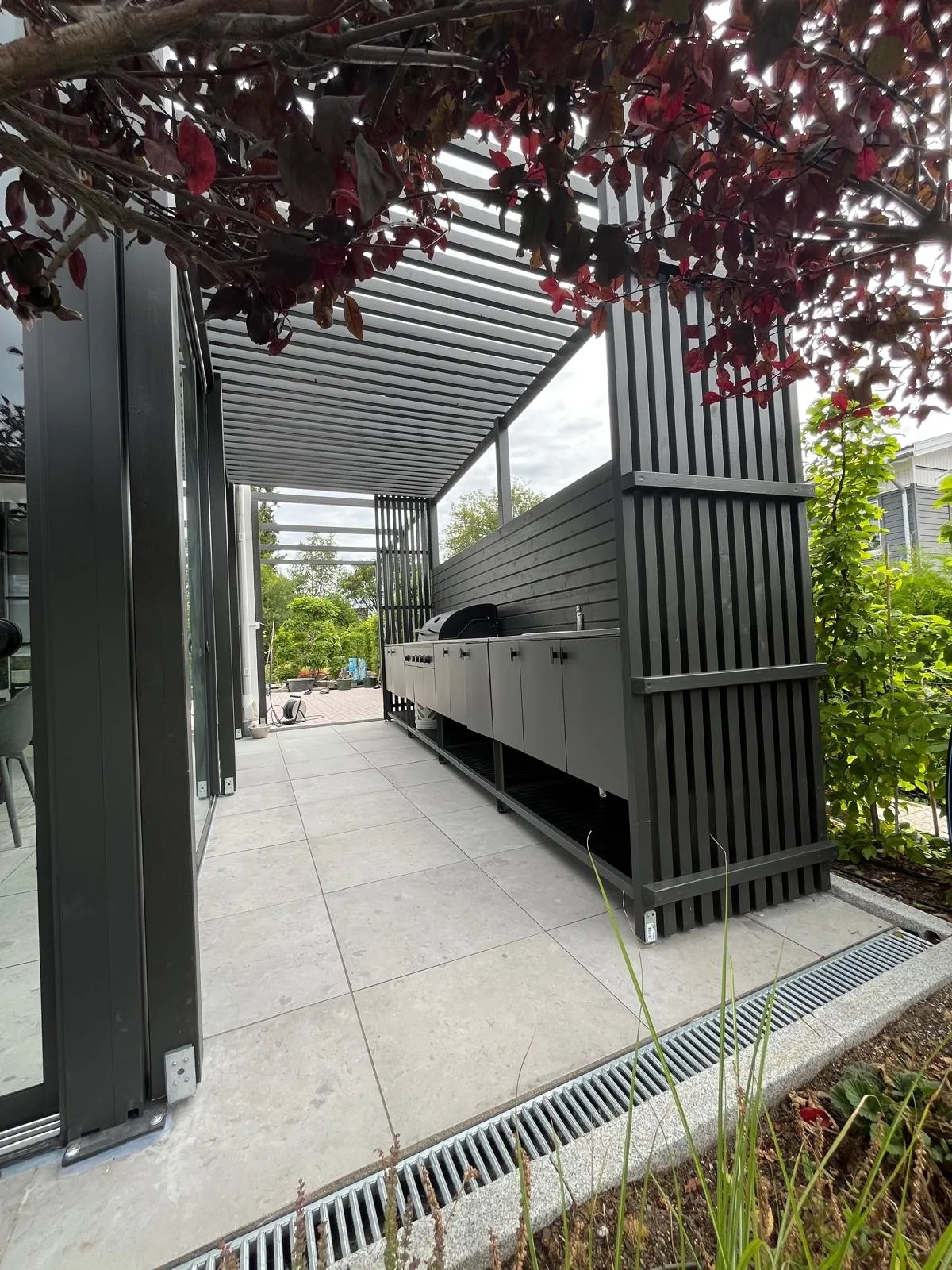 PERGOLUX pergola med ribbtak, insynsskydd och platsbyggt utekök på en stenlagd uteplats.